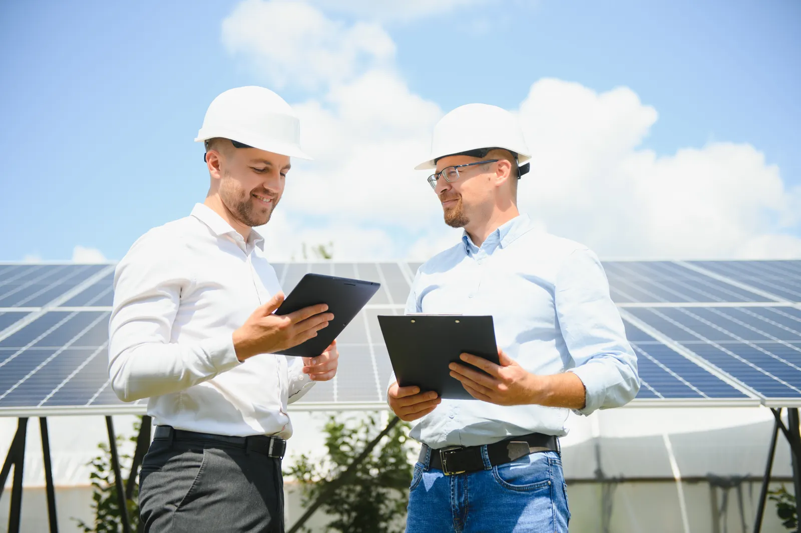Ein Ingenieur und ein Unternehmer mit Schutzhelmen besprechen sich mit Tablet und Klemmbrett vor einer Solaranlage