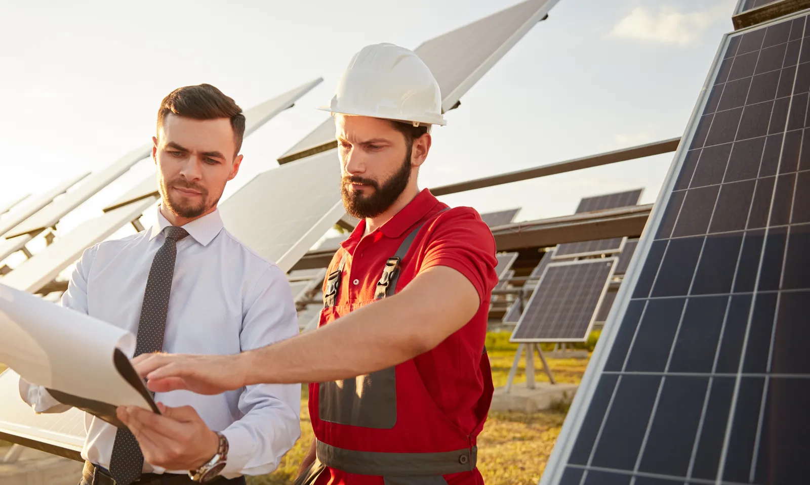 Ein Monteur mit Schutzhelm und ein Mann mit Krawatte gehen Unterlagen zwischen schräg aufgeständerten Solarmodulen durch