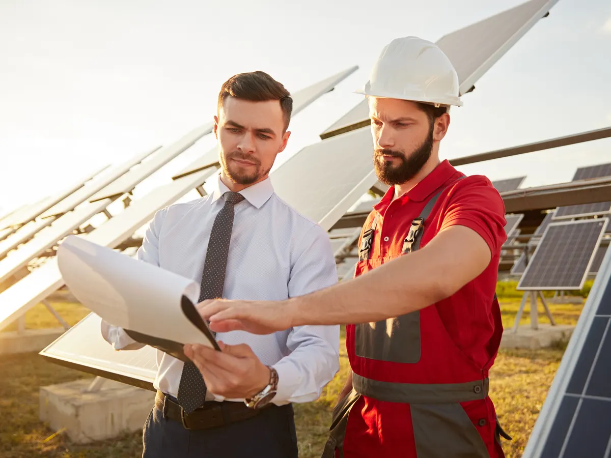 Zwei Personen mit Schutzhelmen besprechen sich auf einer Wiese vor einer Freiflächen-Solaranlage