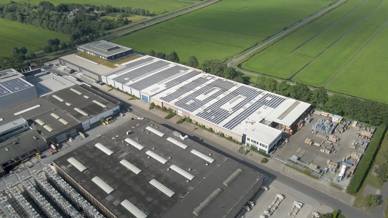 Luftaufnahme eines Gewerbegebiets: lange Industriehalle mit Solarmodulen auf dem Flachdach, daneben Felder