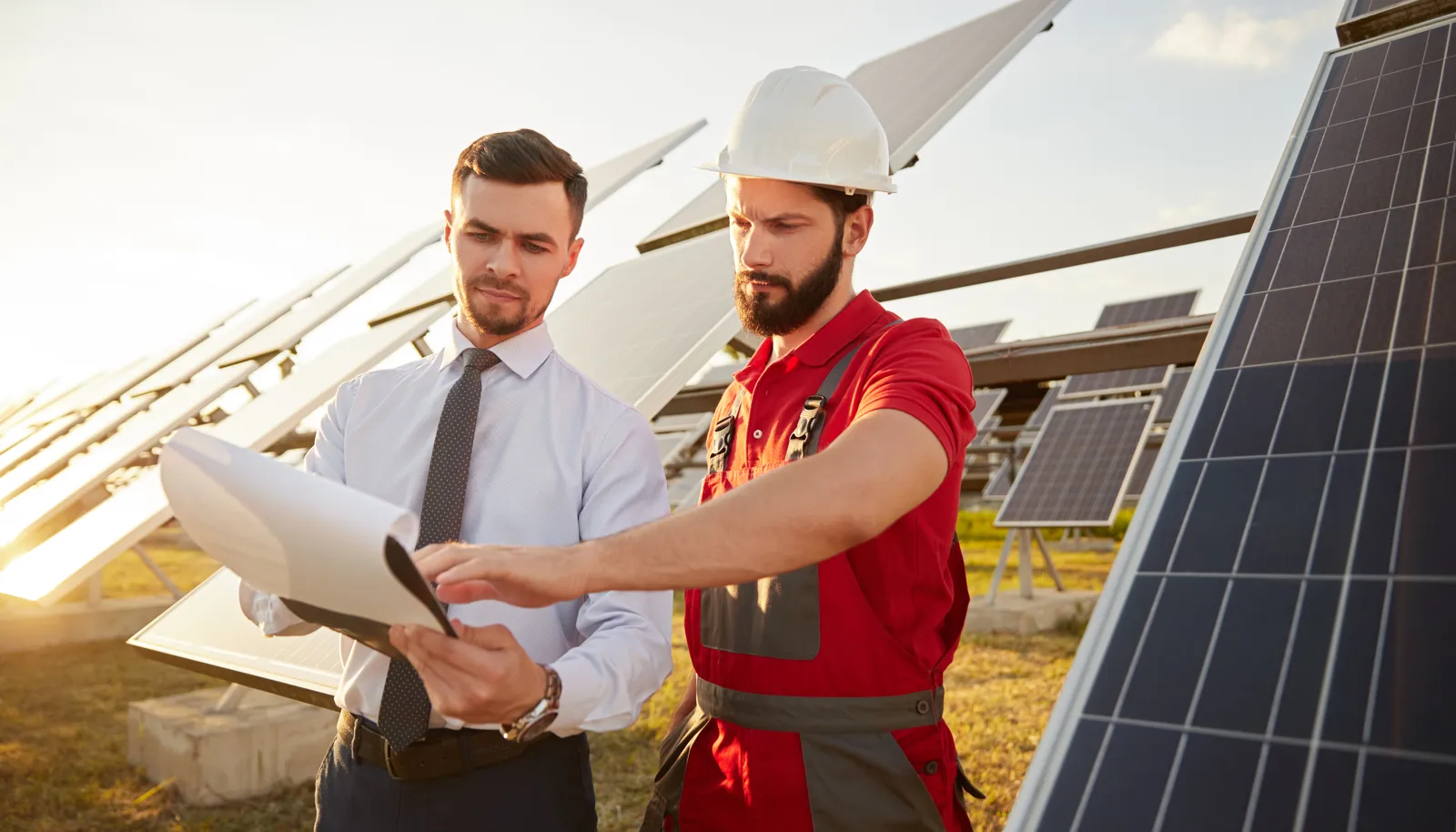 Ein Monteur mit Schutzhelm und ein Mann mit Krawatte prüfen Unterlagen zwischen schräg aufgeständerten Solarmodulen