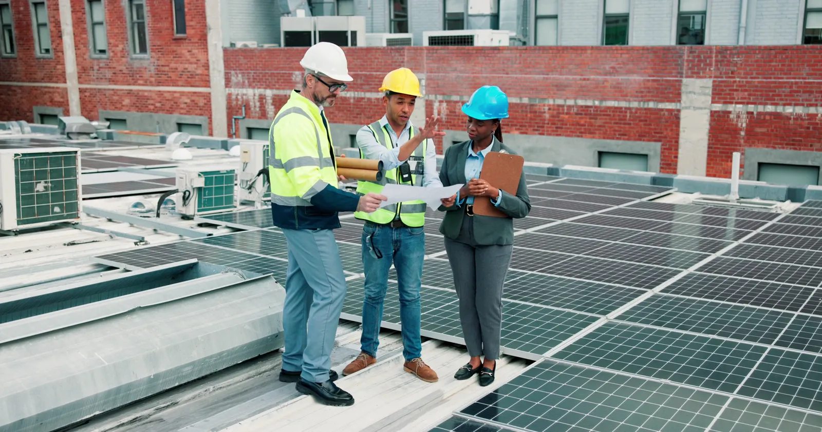 Drei Fachleute mit Schutzhelmen besprechen Unterlagen auf einem Flachdach mit Solarmodulen