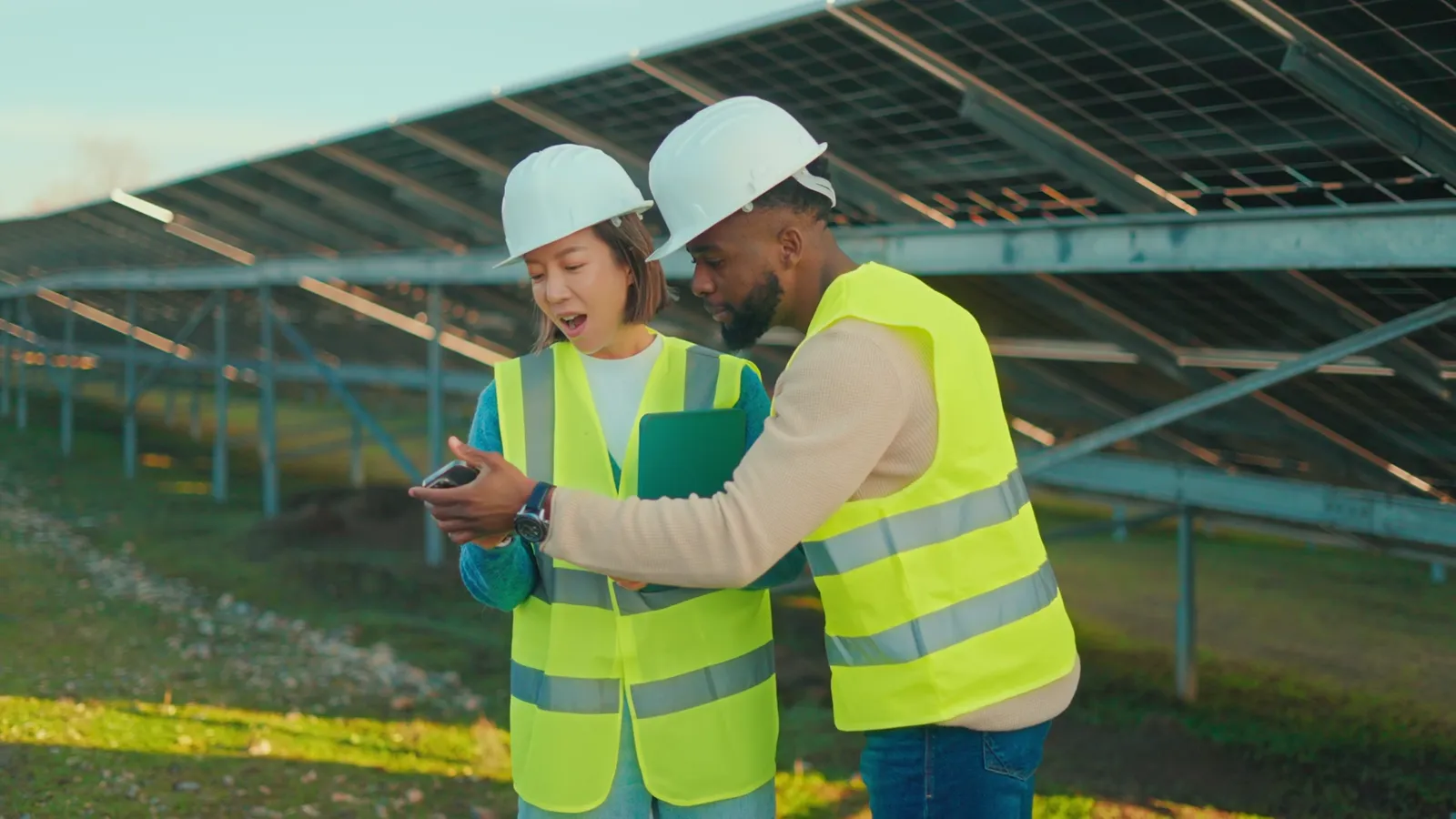 Zwei Fachleute mit Schutzhelmen schauen gemeinsam auf ein Smartphone vor einer Solaranlage