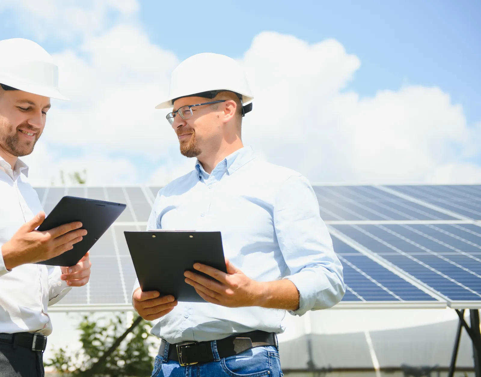 Zwei Männer mit Schutzhelmen besprechen sich mit Tablet und Klemmbrett vor einer Solaranlage