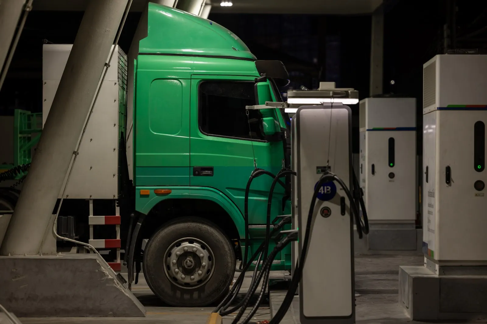 Lkw mit grünem Fahrerhaus an einer DC-Schnellladesäule in einer überdachten Ladestation, daneben weitere Ladeschränke
