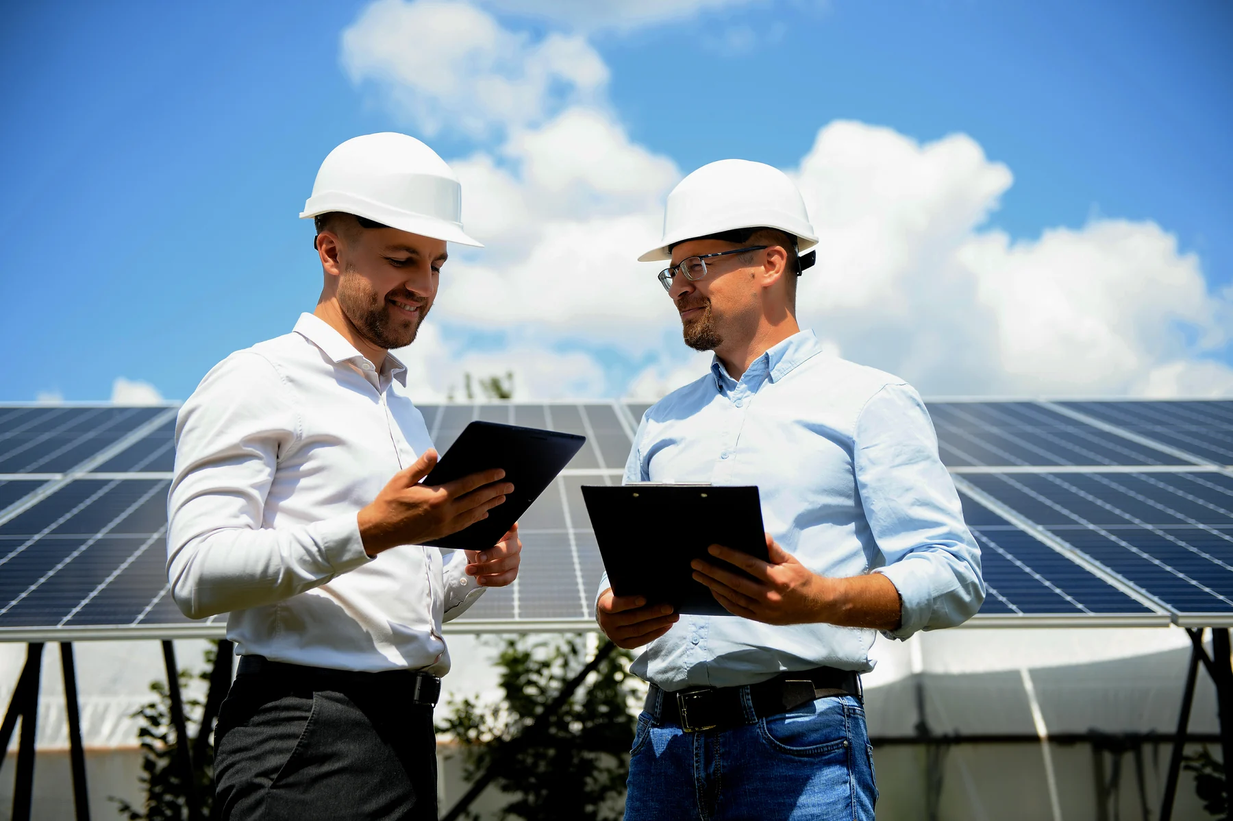 Ein Unternehmer und ein Ingenieur mit Schutzhelmen besprechen sich mit Tablet und Klemmbrett vor einer großen Solaranlage, sonniger Tag